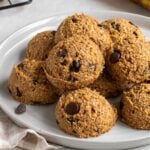 Close up of a white speckled plate of banana bread bites with dark chocolate chips on the side, a banana in the background, and a beige checkered pattern tea towel on the side.