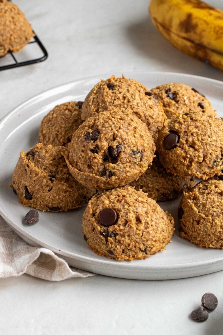 Close up of a white speckled plate of banana bread bites with dark chocolate chips on the side, a banana in the background, and a beige checkered pattern tea towel on the side.