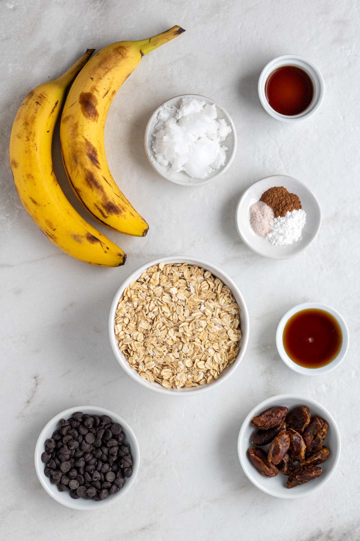 Two ripe bananas with small bowls of vanilla extract, coconut oil, cinnamon, baking powder, salt, rolled oats, maple syrup, dates, and dark chocolate chips on a white background.