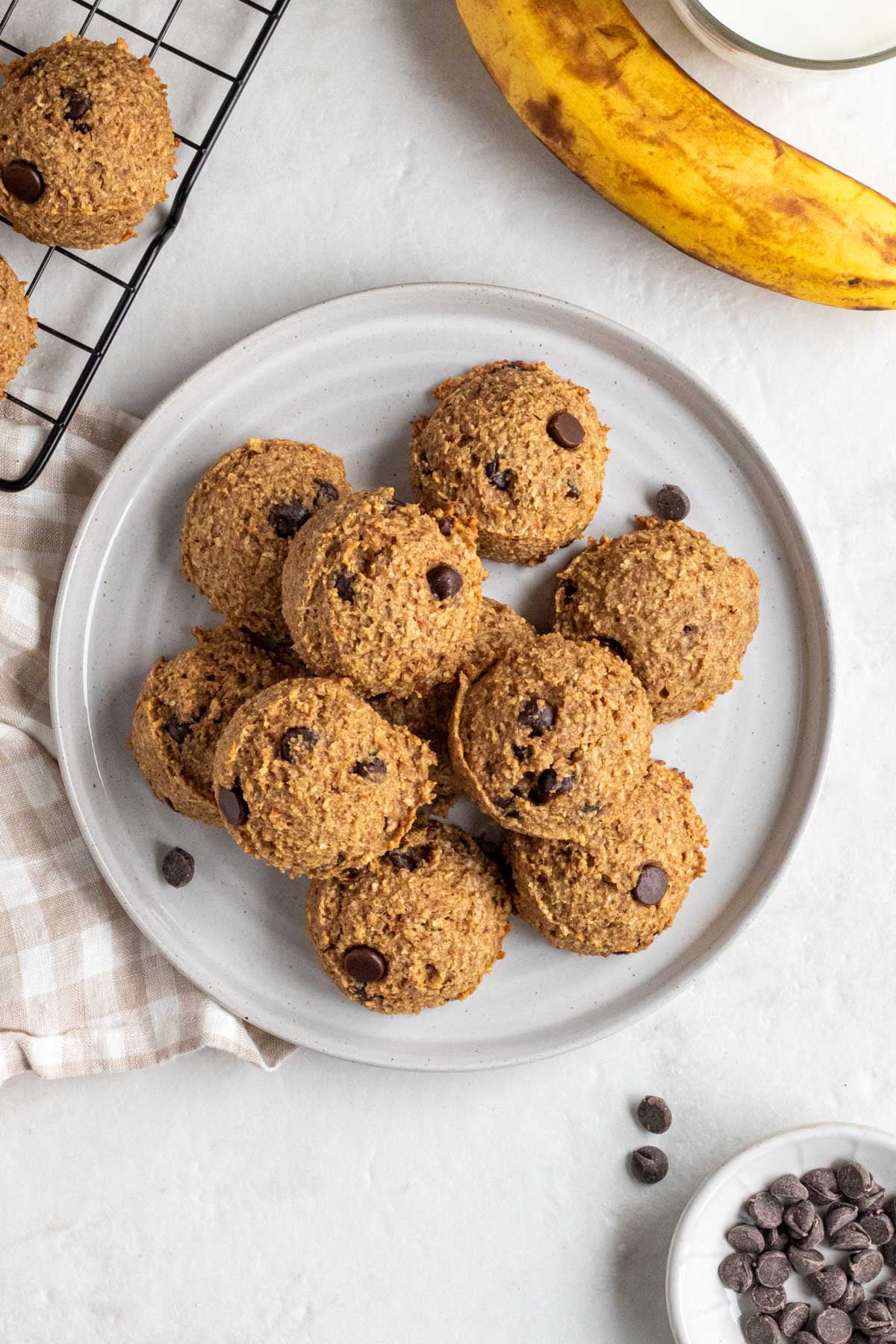A plate of banana bread bites with a wire rack of more bites, glass of milk, a ripe banana, small bowl of dark chocolate chips, and beige and white tea towel on the side on a white background.