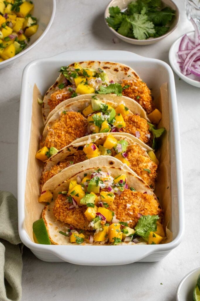 White baking dish filled with coconut crusted fish tacos with avocado mango salsa with a bowl of cilantro and sliced red onion on the side on a white background.