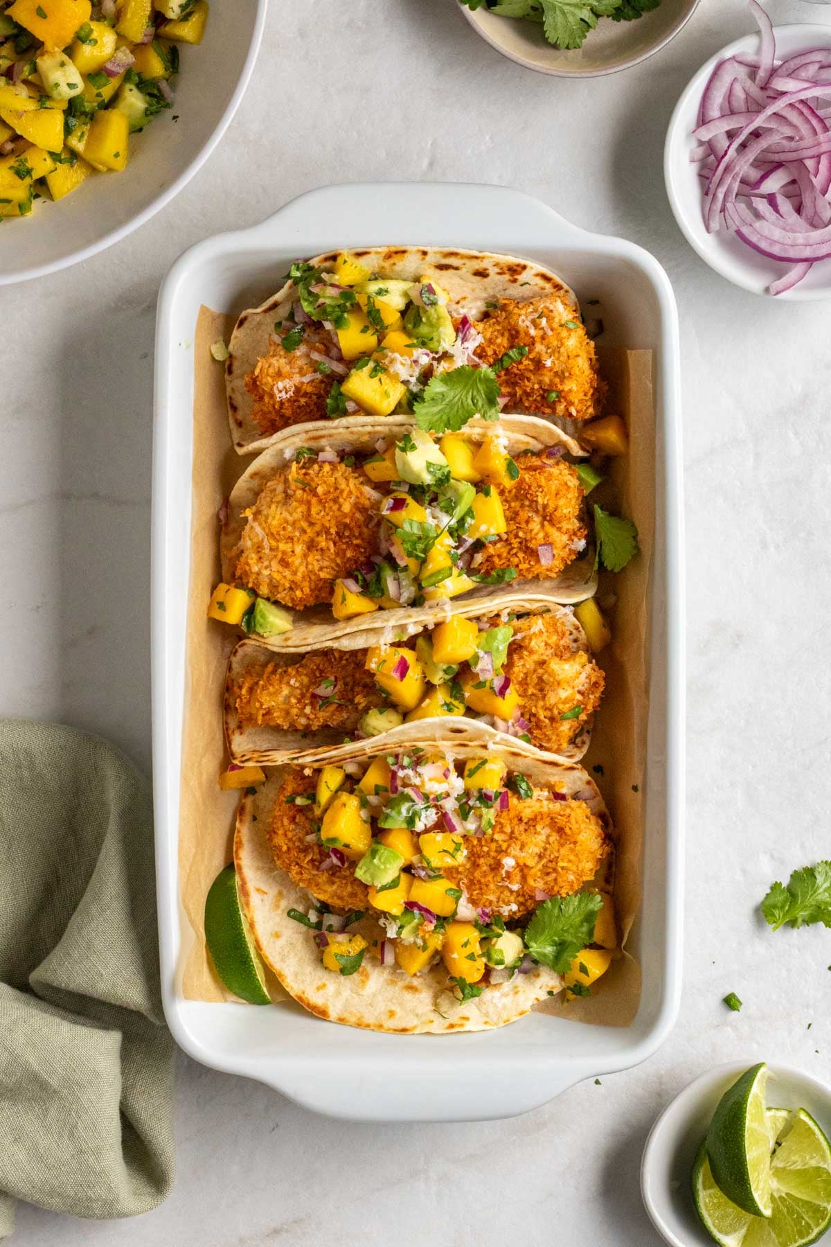 Baking dish filled with coconut crusted fish tacos with mango avocado salsa, fresh cilantro, sliced red onion, and lime wedges with a green tea towel on a white background.