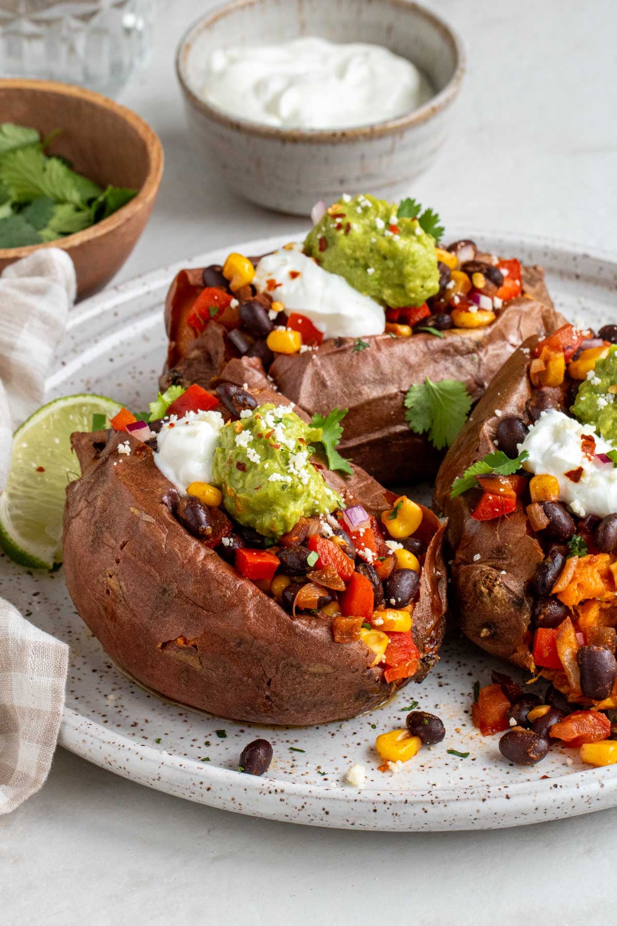 Three baked sweet potatoes stuffed with a black bean filling topped with sour cream and avocado on a white plate with lime wedges on the side.