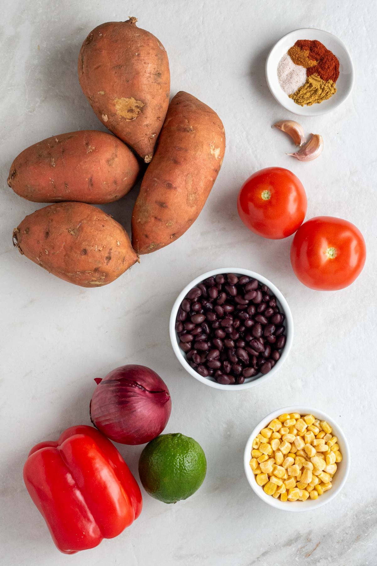 Sweet potatoes, bowl of spices, tomatoes, bowl of black beans, red bell pepper, red onion, lime, and bowl of frozen corn on a white background.