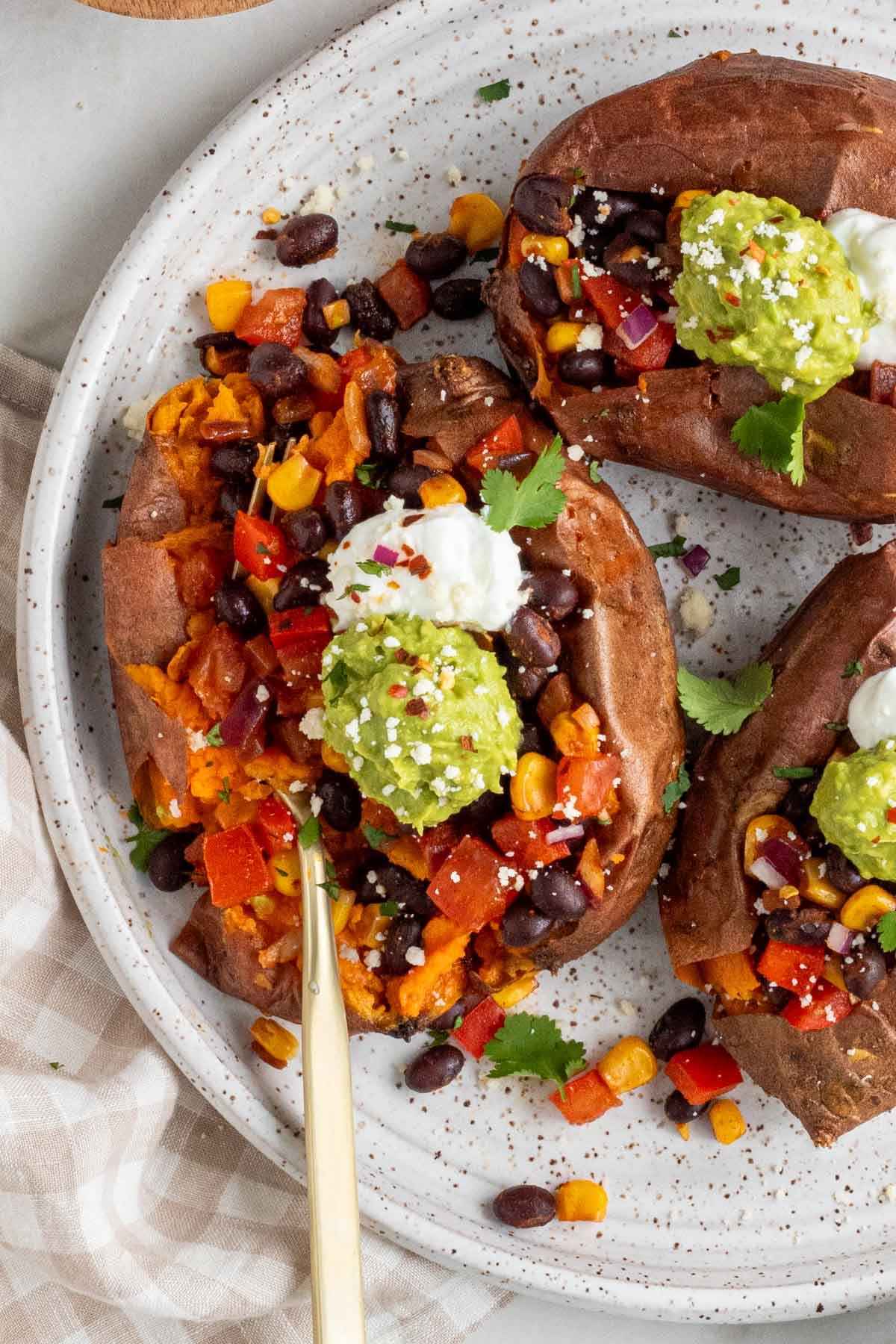 Close up of three black beans stuffed sweet potatoes on a white speckled plate with mashed avocado, sour cream, and fresh cilantro on top with a gold fork and a checkered beige and white tea towel on the side.
