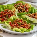 Four Asian chicken lettuce wraps on a plate with a bowl of sauce in the background and a bowl of sesame seeds on the side.