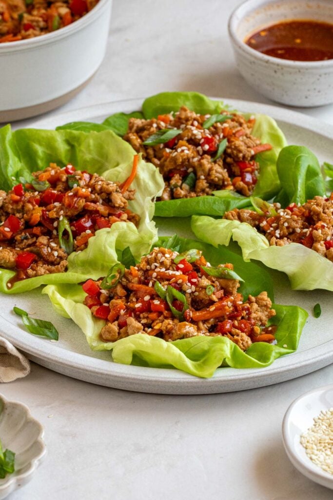 Four Asian chicken lettuce wraps on a plate with a bowl of sauce in the background and a bowl of sesame seeds on the side.