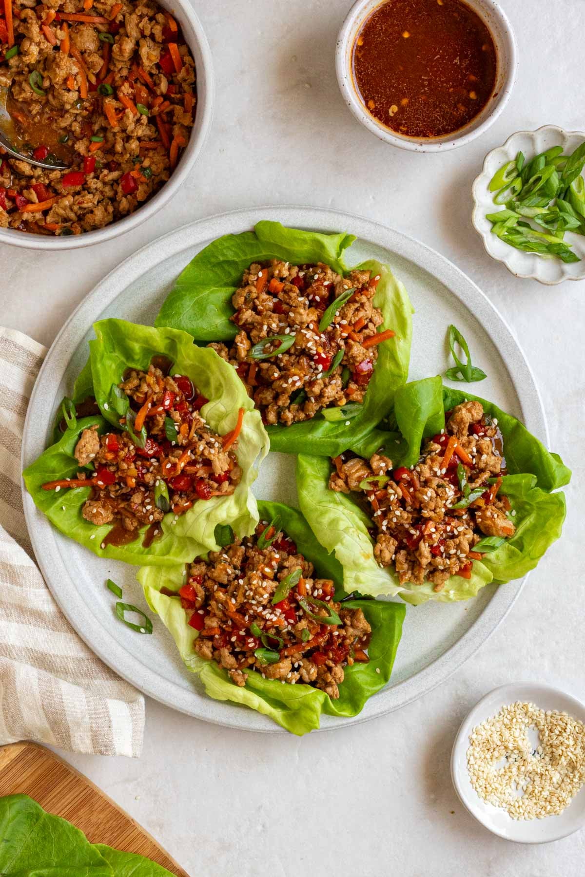 Asian chicken lettuce wraps on a plate with extra ground meat mixture, sauce, sesame seeds, and lettuce on the side.