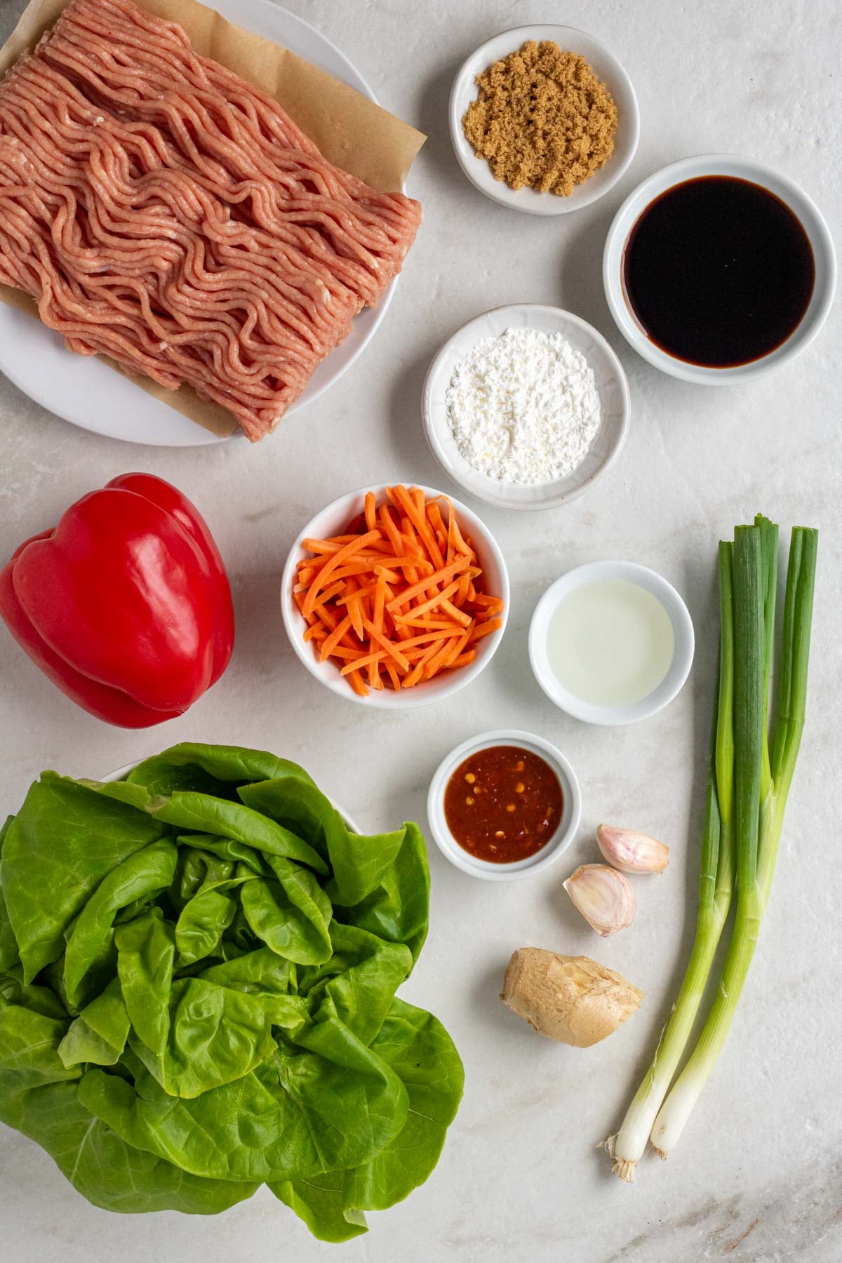 Plate of ground chicken with bowls of brown sugar, soy sauce, corn starch, rice vinegar, shredded carrots, and hot sauce on teh side with a red bell pepper, head of lettuce, green onions, garlic, and ginger root on the side on a white background.