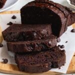 Chocolate zucchini bread on a wood cutting board on a piece of parchment paper with dark chocolate chips on the side and a glass of milk in the background.