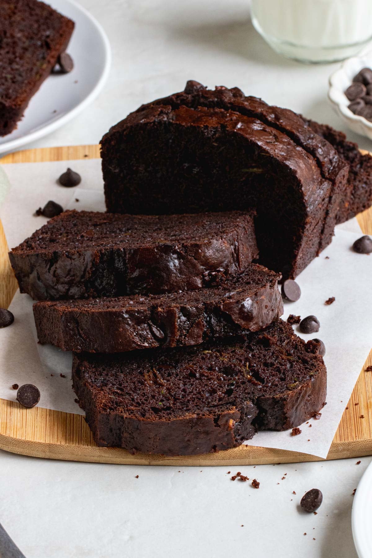 Chocolate zucchini bread on a wood cutting board on a piece of parchment paper with dark chocolate chips on the side and a glass of milk in the background.