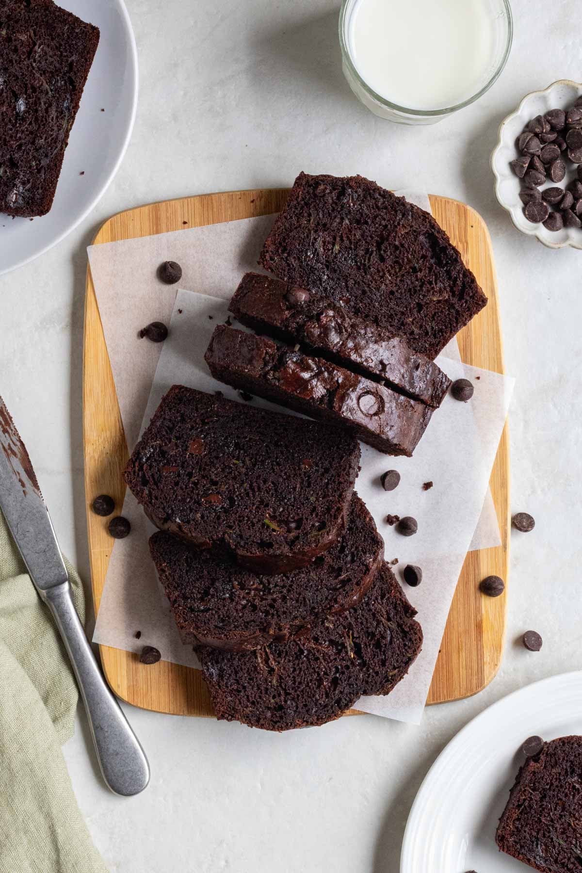 Slices of double chocolate zucchini bread on a wood cutting board with slices on plates on the side with dark chocolate chips, glass of milk, knife, and light green tea towel on a white background.