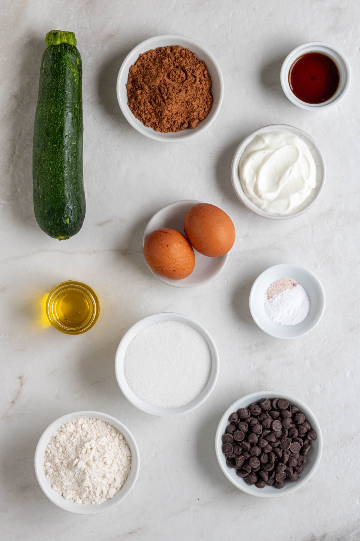 Small zucchini, bowl of cocoa powder, bowl of vanilla extract, bowl of Greek yogurt, 2 eggs, jar of olive oil, bowl of baking soda and salt, bowl of sugar, bowl of flour, and bowl of dark chocolate chips on a white background.