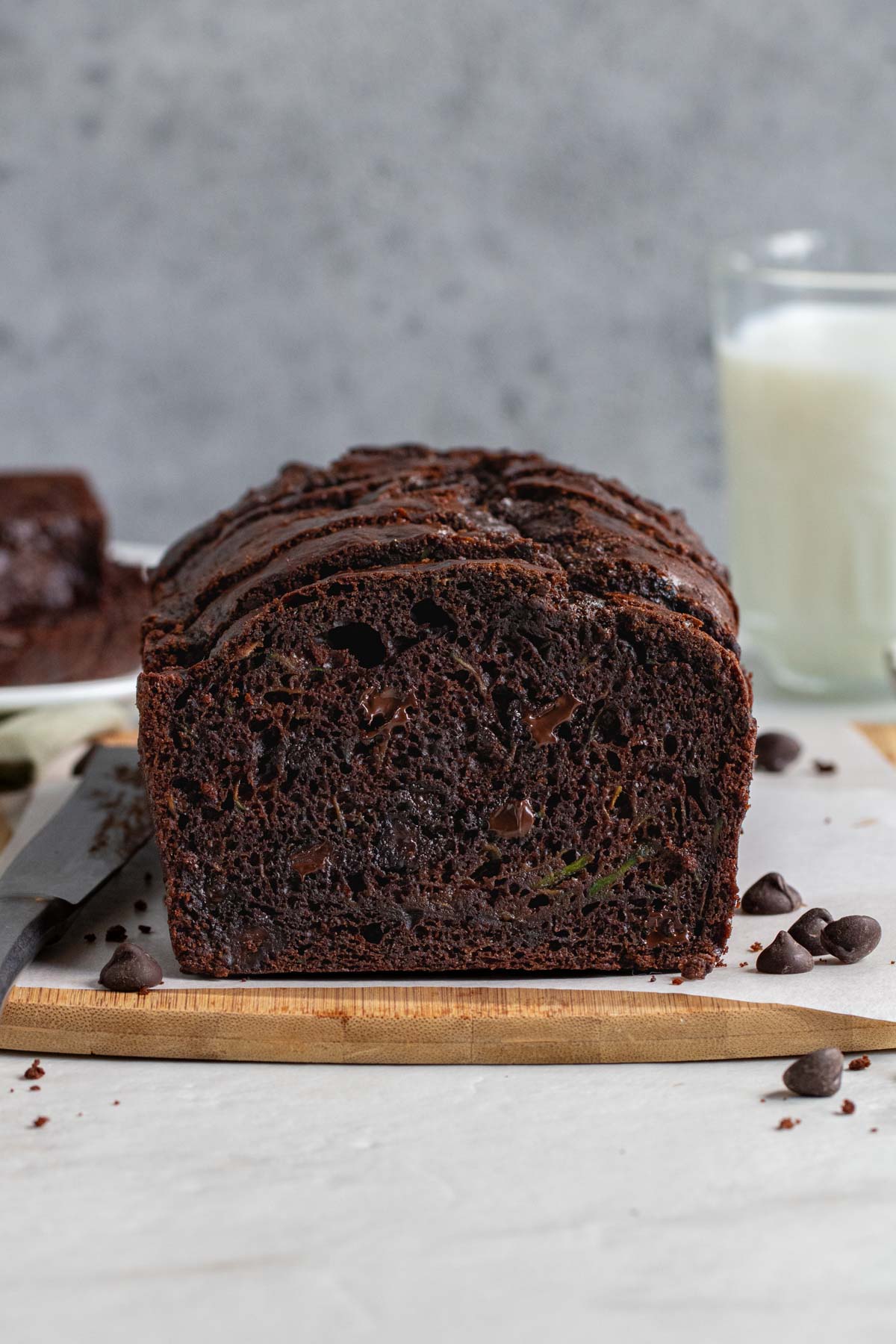 Close of up a loaf of chocolate zucchini bread with a glass of milk and chocolate chips on the side on a dark blue background.