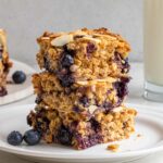 Three blueberry baked oatmeal bars stacked on a white plate with a glass of milk in the background and fresh blueberries on the side on a white background.