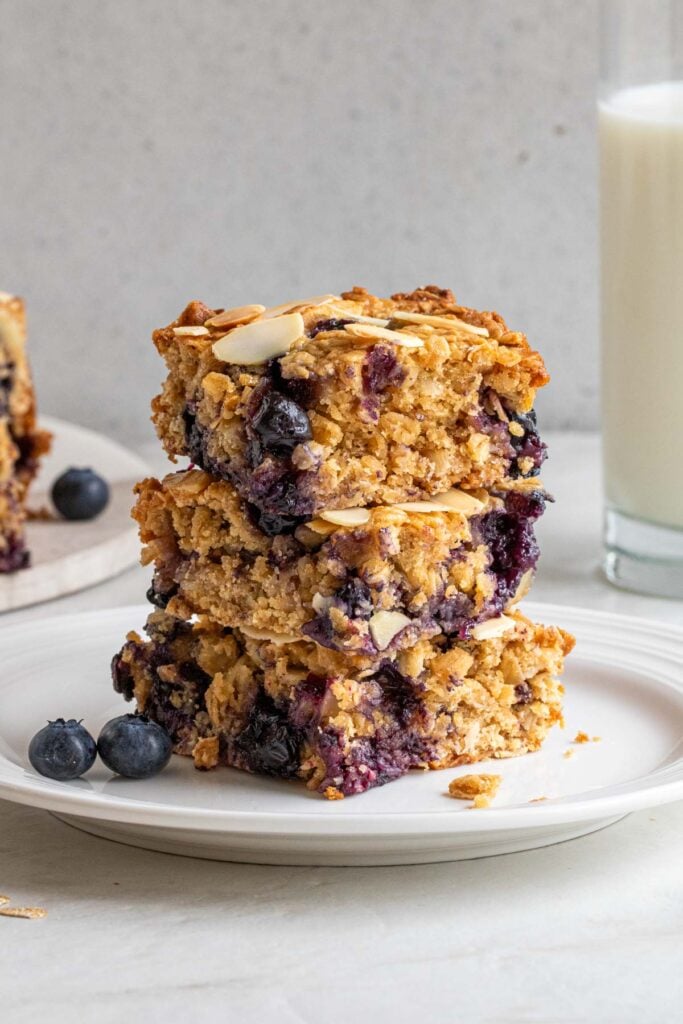 Three blueberry baked oatmeal bars stacked on a white plate with a glass of milk in the background and fresh blueberries on the side on a white background.