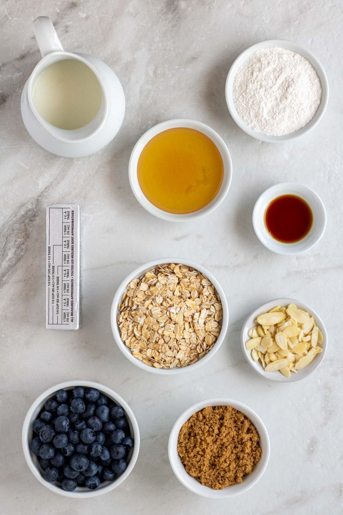 Milk, honey, all-purpose flour, vanilla extract, rolled oats, slivered almonds, brown sugar, and fresh blueberries in a bowl with a stick of butter on the side on a white background.