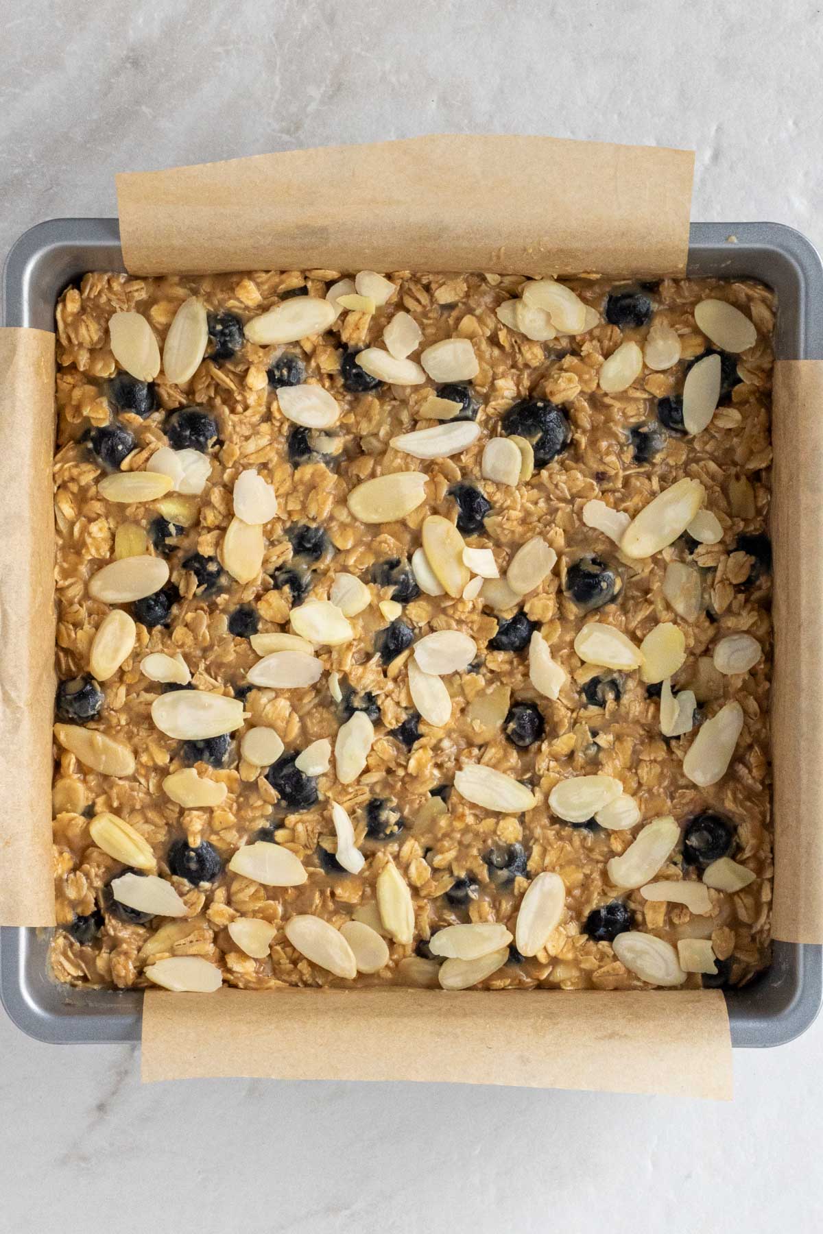 Raw blueberry baked oatmeal batter in a silver baking pan with parchment paper on a white background.