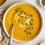 Bowl of roasted pumpkin soup with cream, pumpkin seeds, and parsley on top on a plate with a spoon with a checkered tea towel on the side with crusty bread, a bowl of salt, and a jar of cream on the side.