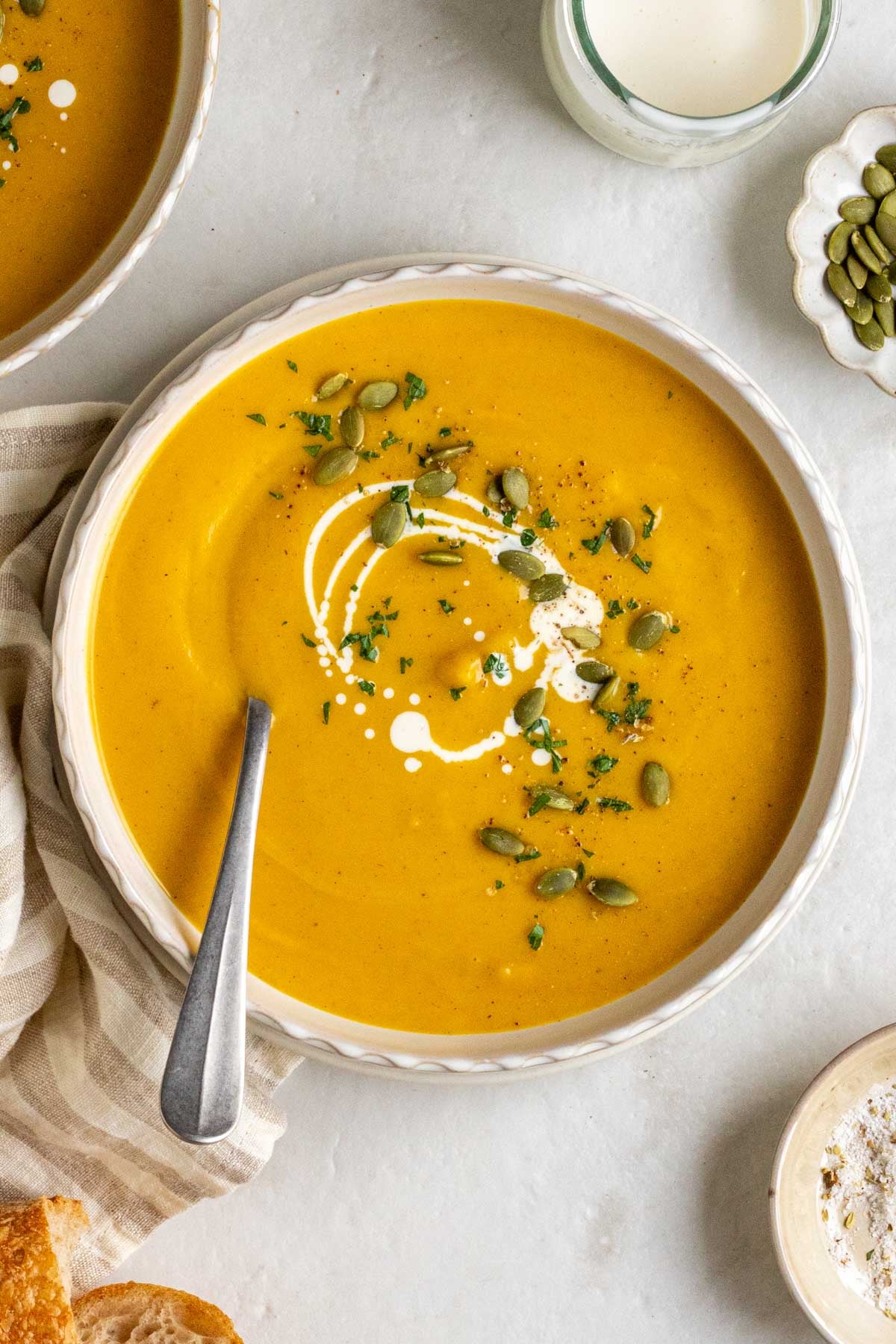 Bowl of roasted pumpkin soup with cream, pumpkin seeds, and parsley on top on a plate with a spoon with a checkered tea towel on the side with crusty bread, a bowl of salt, and a jar of cream on the side. 