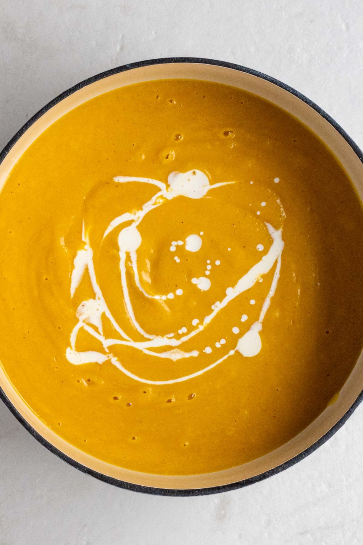 Large pot creamy pumpkin soup with heavy cream drizzled on top on a white background.