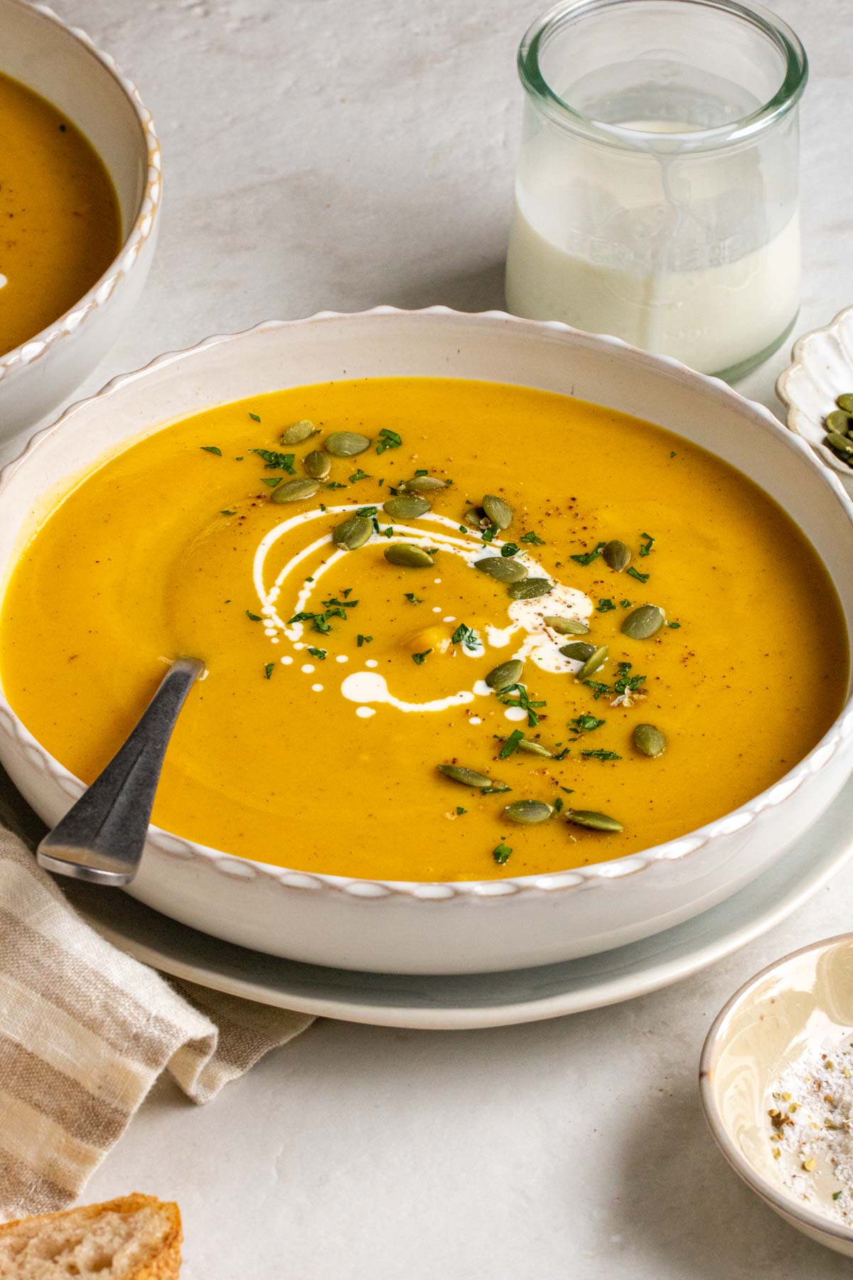 A close up of a bowl of creamy pumpkin soup with a drizzle of heavy cream, pumpkin seeds, and chopped parsley on top.