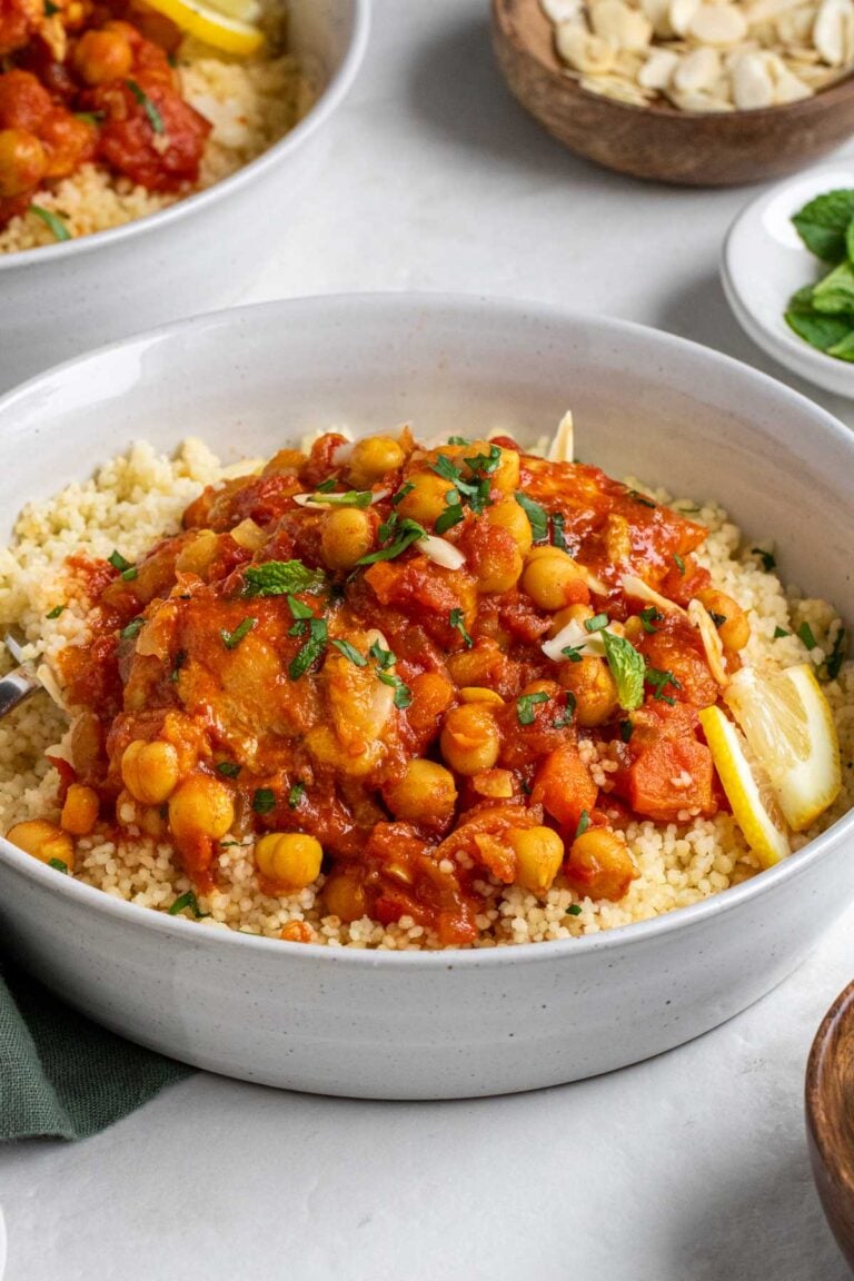 Bowl of slow cooker moroccan chicken served over couscous with fresh cilantro and fresh mint on top with a fork on a white background.