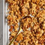 Close up of vanilla almond granola on gold serving spoon on a silver baking sheet with parchment paper on a white background.