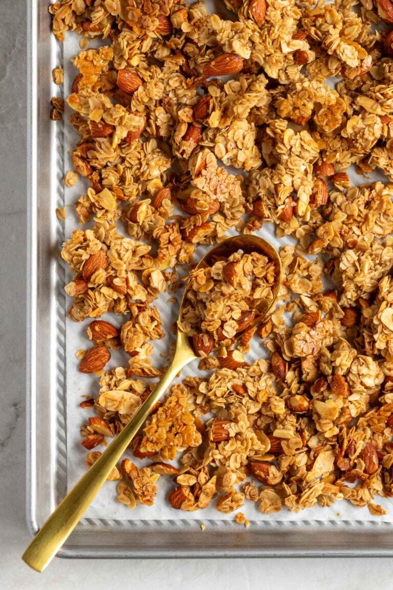 Close up of vanilla almond granola on gold serving spoon on a silver baking sheet with parchment paper on a white background.