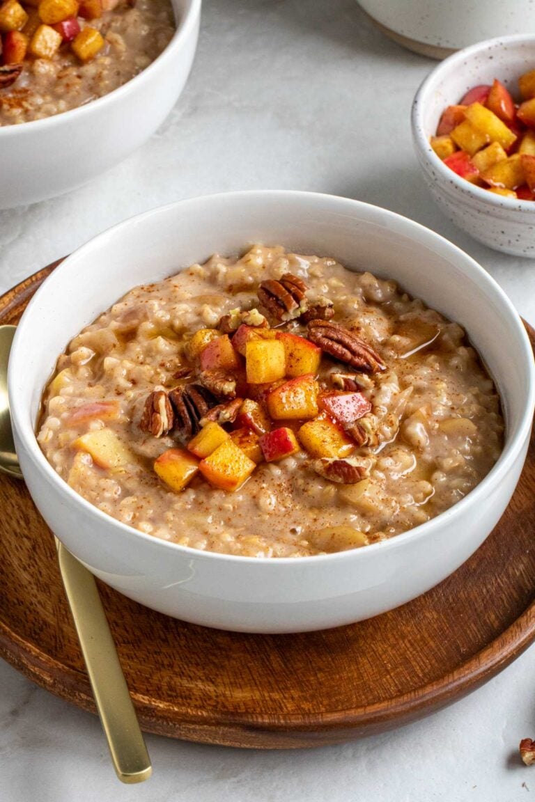 White bowl of slow-cooker apple cinnamon oatmeal with sautéed apples, pecans, and maple syrup on top on a dark wood plate with a gold spoon.