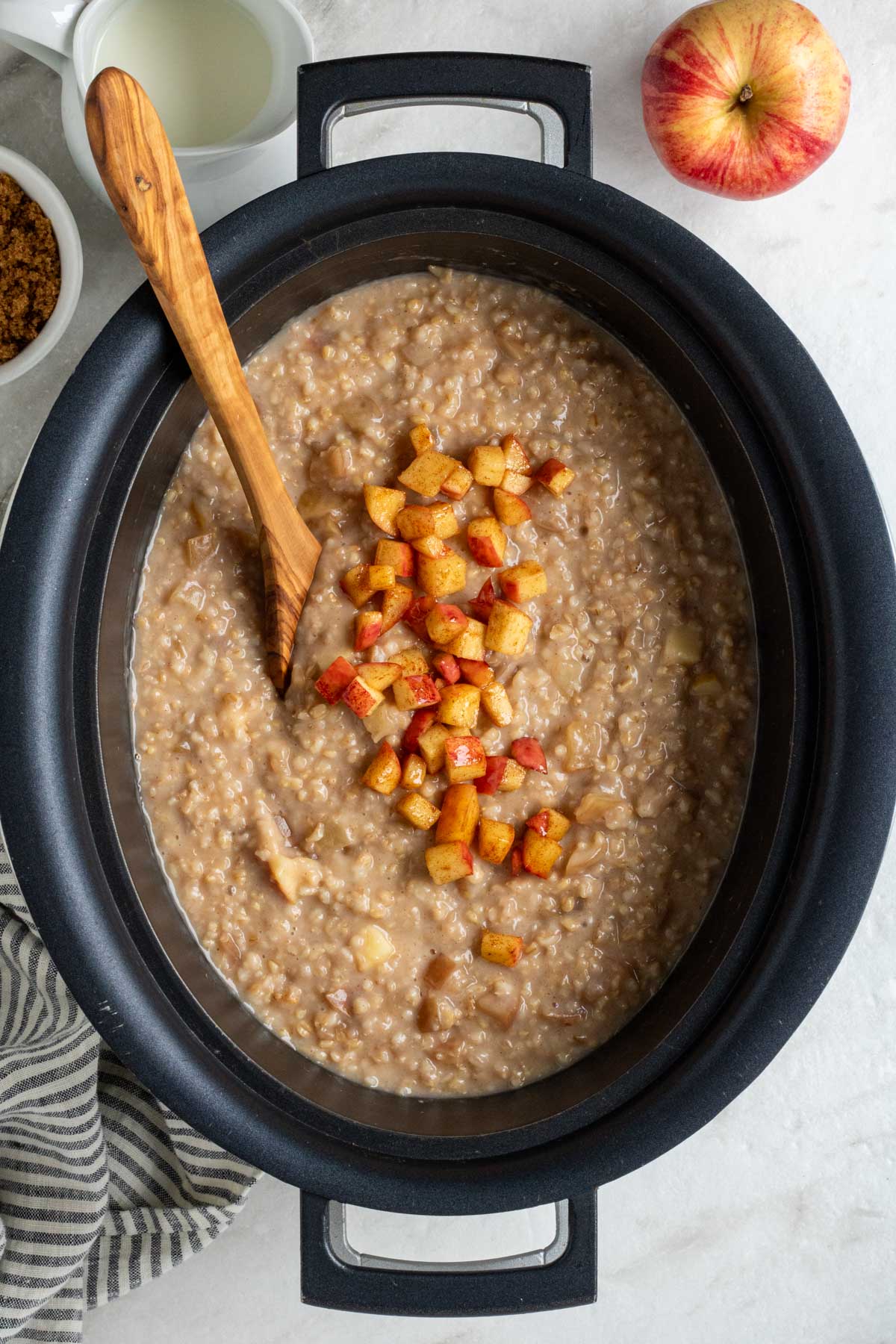 A crockpot filled with cooked slow cooker apple cinnamon oatmeal with sautéed apples on top with a wood serving spoon with milk, brown sugar, fresh apple, and tea towel on the side on a wide background.