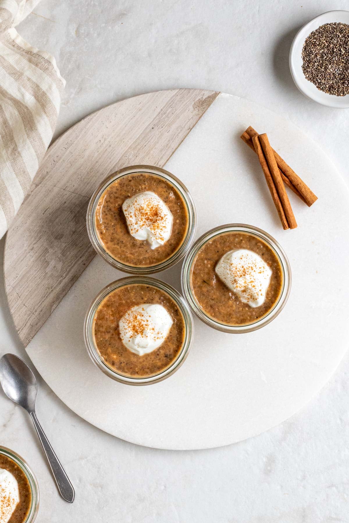 Three jars pumpkin chia seed pudding with cinnamon sticks on a white cutting board with a small silver spoon and a bowl of chia seeds on the side on a white background.
