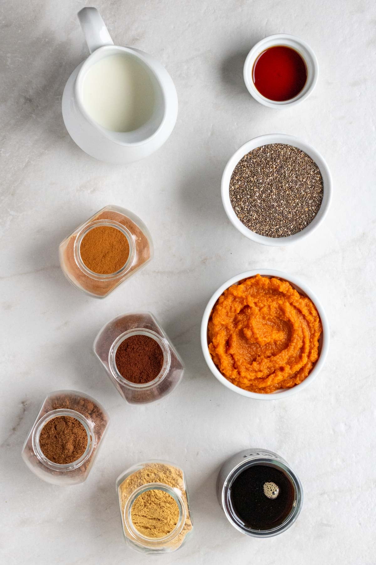 Jug of milk, bowls of maple syrup, chia seeds, pumpkin puree, jars of cinnamon, cloves, nutmeg, and ginger, and a jar of maple syrup on a white background.