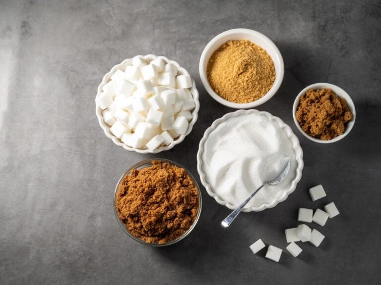 Bowls of sugar brown, brown sugar, demerara sugar, raw sugar, and white sugar on a dark grey background.