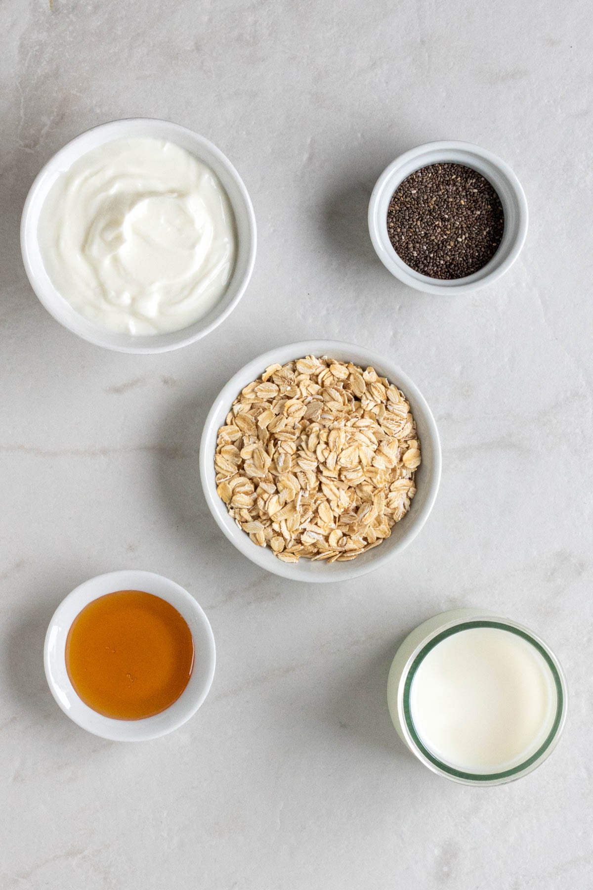 Bowl of Greek yogurt, bowl of chia seeds, bowl of rolled oats, bowl of honey, and jar of milk on a white background.