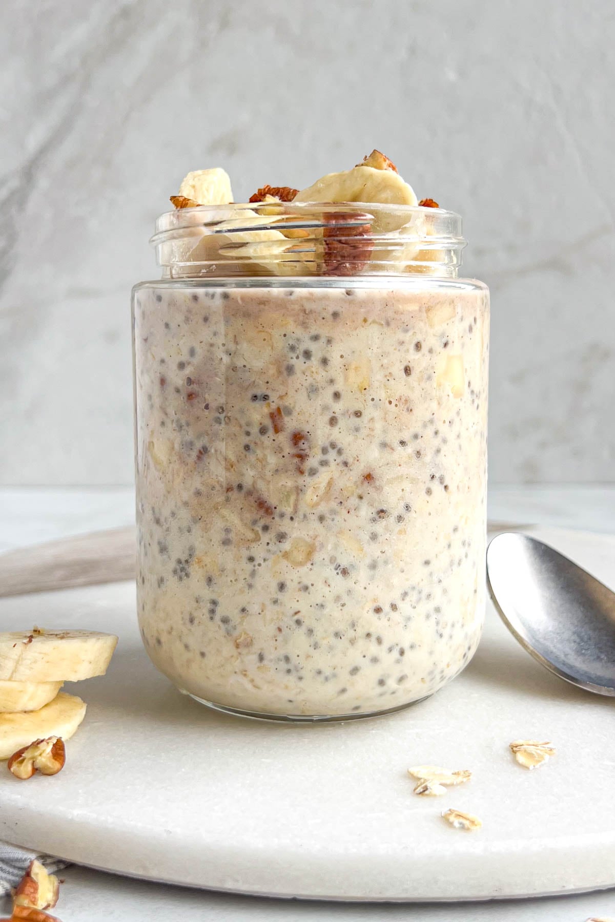 Jar of banana bread overnight oats on a white background with a spoon, rolled oats, and sliced banana and pecans on the side.