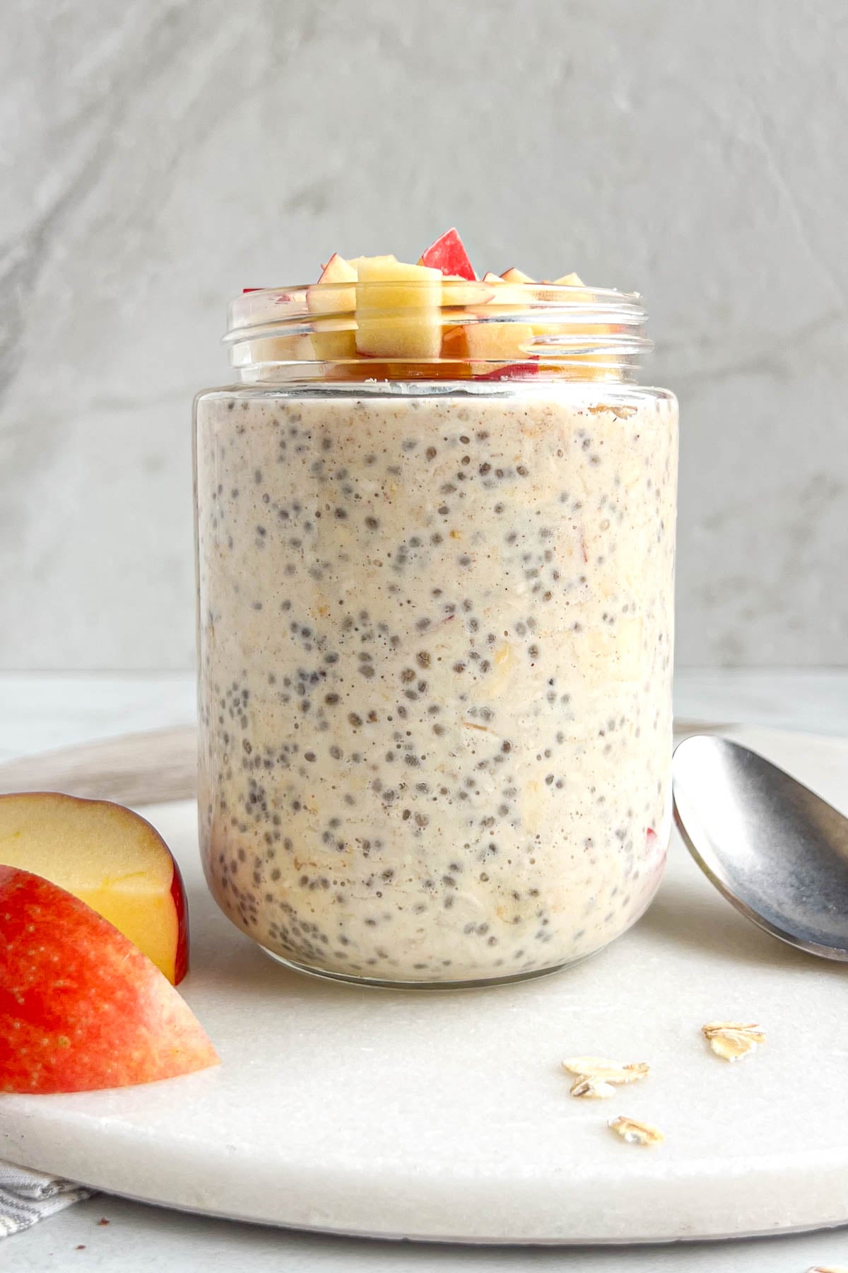 Jar of apple pie overnight oats on a white background with a spoon, rolled oats, and apple slices on the side.