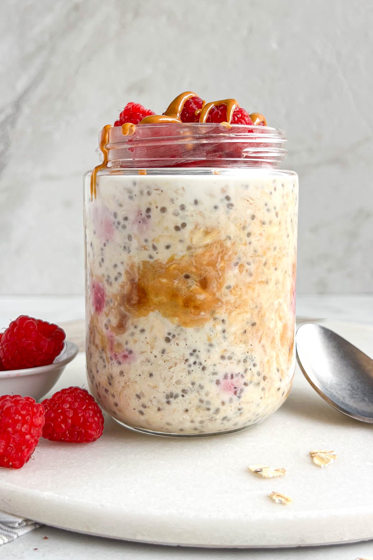 Jar of peanut butter and jelly overnight oats on a white background with a spoon, rolled oats, and fresh raspberries on the side.