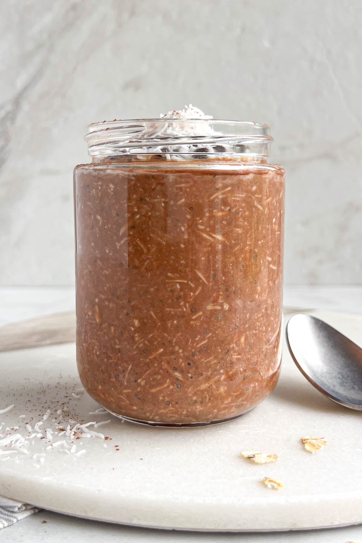 Jar of chocolate coconut overnight oats on a white background with a spoon, rolled oats, and shredded coconut on the side.