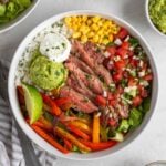 Two steak fajita bowls with a grey and white striped tea towel with a silver fork and bowls of pico de gallo, guacamole, sour cream, and cilantro on the side.