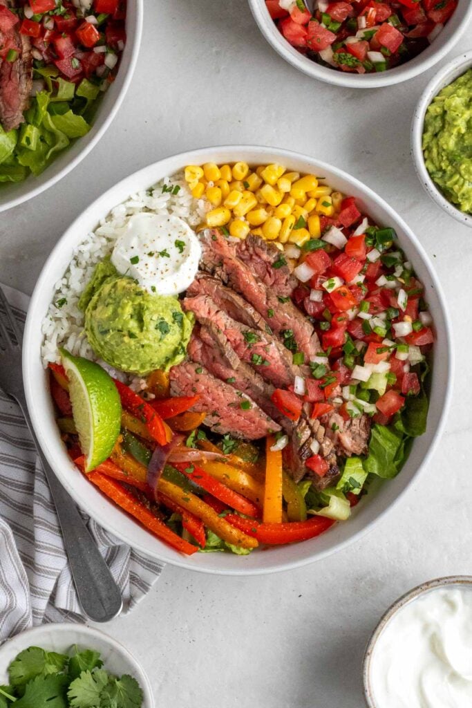 Two steak fajita bowls with a grey and white striped tea towel with a silver fork and bowls of pico de gallo, guacamole, sour cream, and cilantro on the side.
