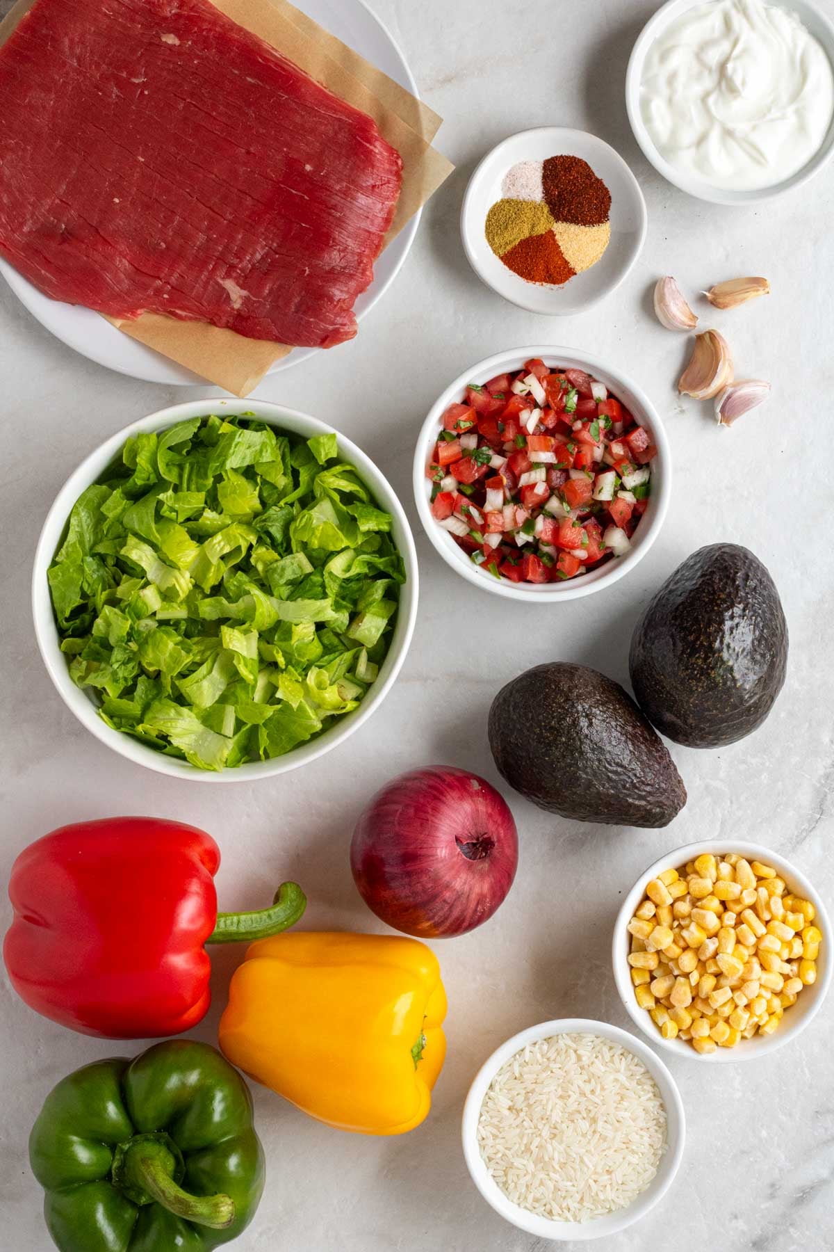 Plate of flank steak with a bowl of spices, pico de gallo, shredded lettuce, frozen corn, white rice, garlic, avocados, red onion, and bell peppers on a white background.