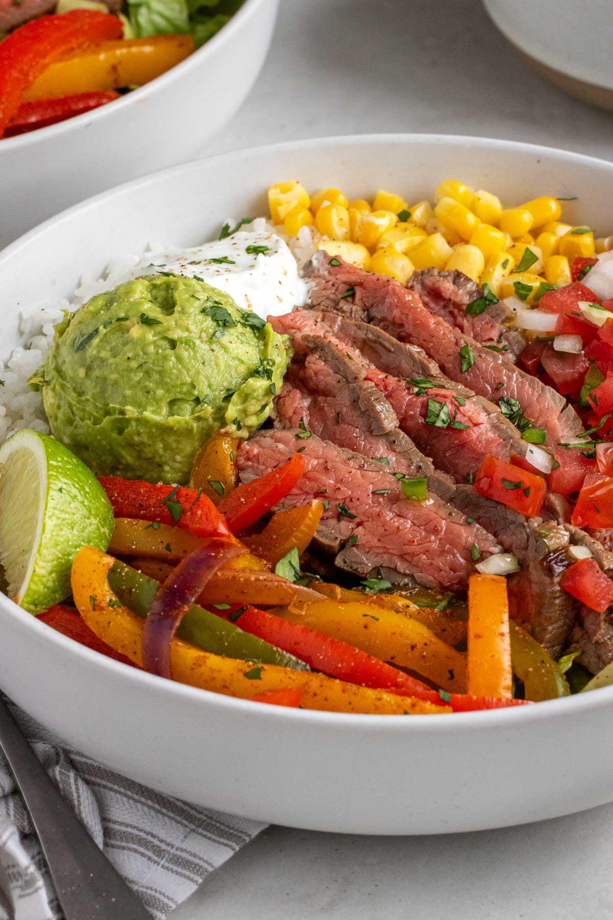 Close up of a white bowl with cooked rice, sliced flank steak, fajita veggies, corn, pico de gallo, guacamole, sour cream, and a lime wedge.