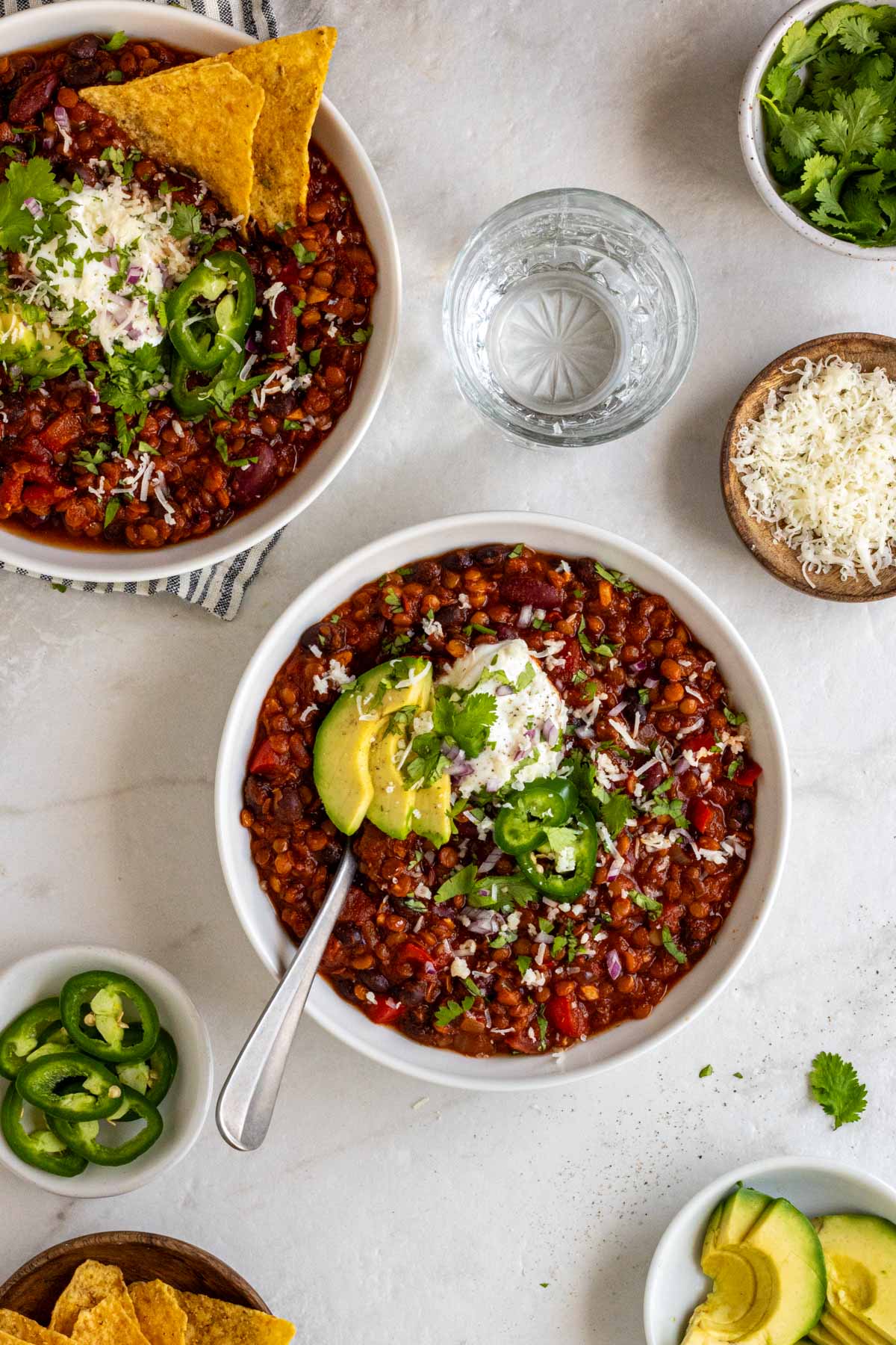 Two bowls of lentil chili with fresh cilantro, shredded cheese, sliced jalapeño, and slice avocado on a white background.