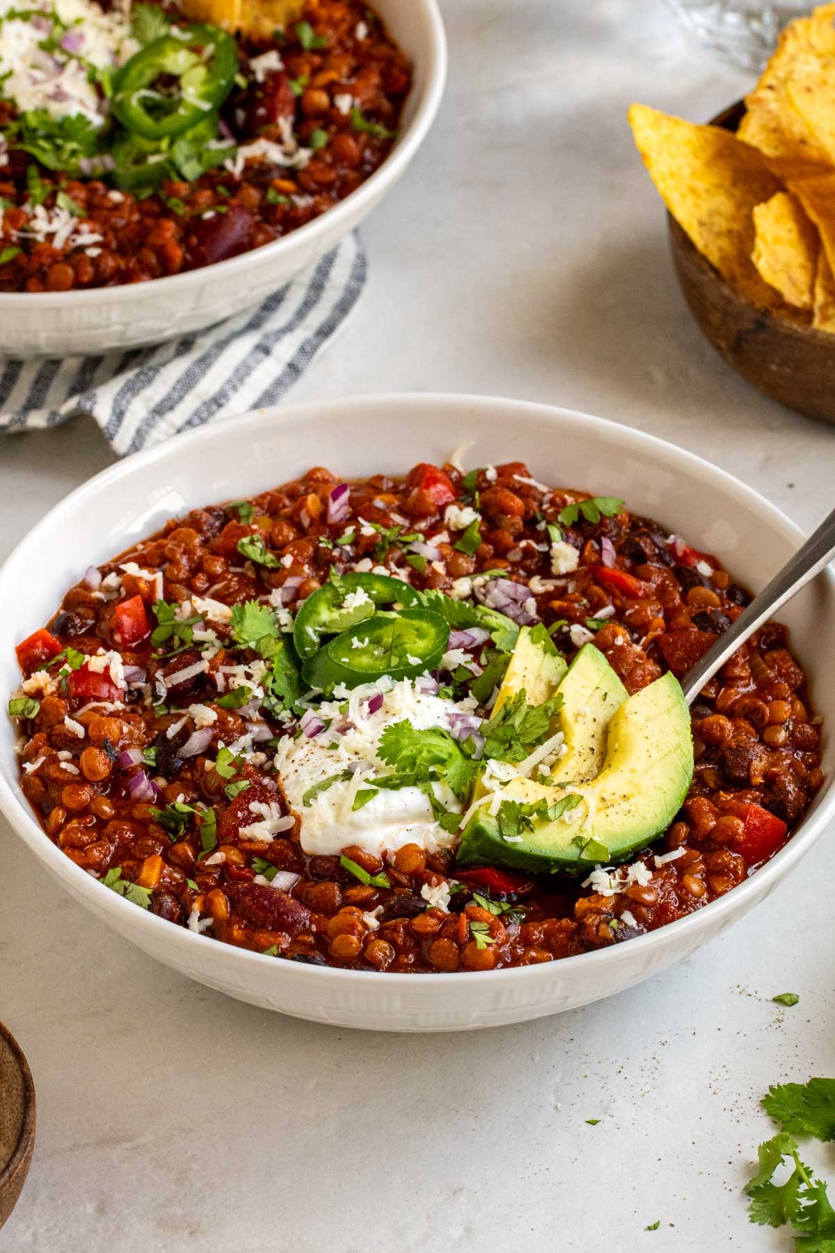 Close up of two bowls of vegetarian lentil chili with a bowl of shredded cheese and tortilla chips on the side on a white background..