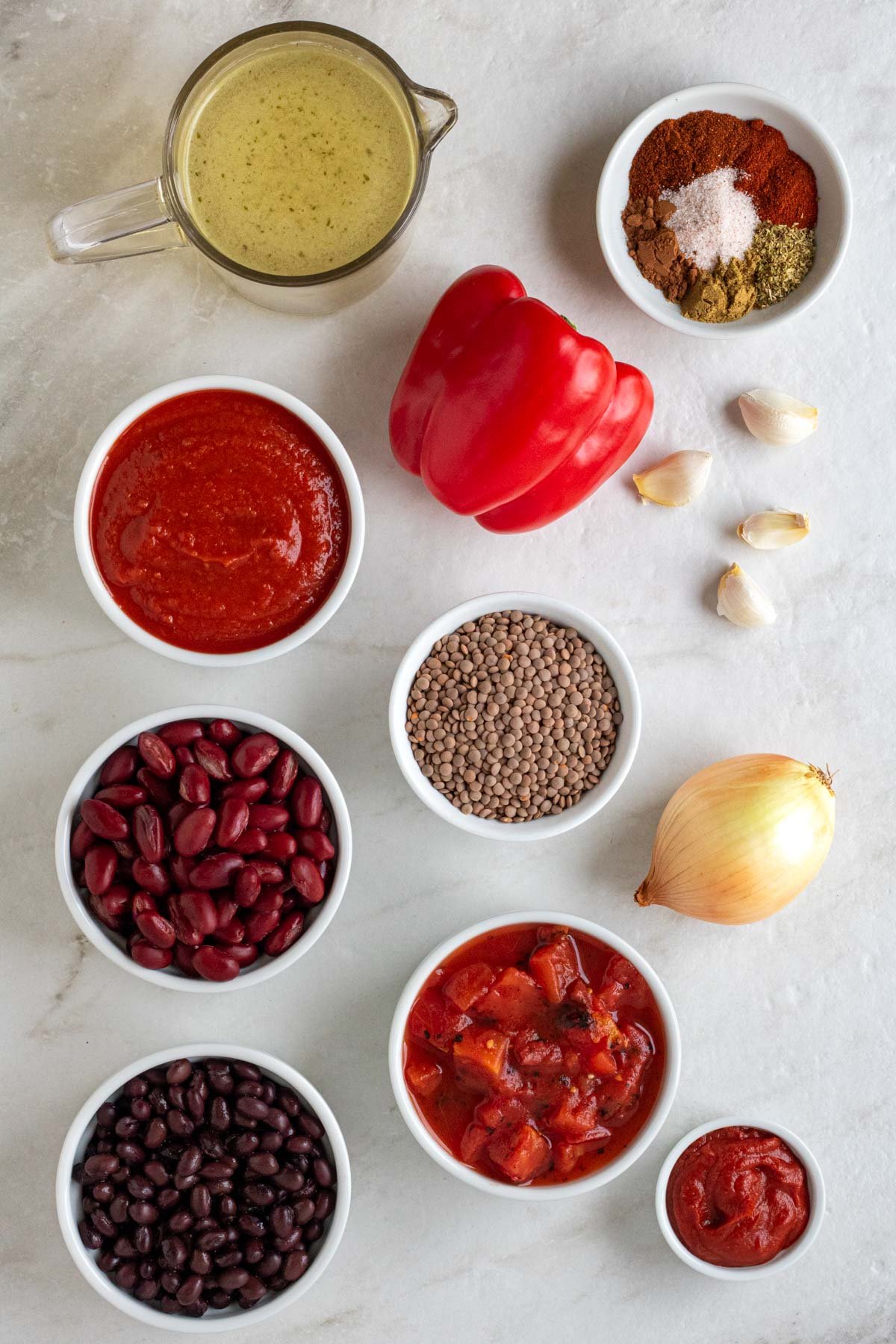 Bowls of brown lentils, red kidney beans, black beans, diced tomatoes, crushed tomatoes, tomato paste, and spices, with a yellow onion, garlic cloves, and a jug of vegetable broth on a white background. 