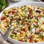 Large white bowl of mediterranean orzo salad with a silver serving spoon on a beige and white checkered tea towel with bowls of parsley, feta cheese, and olives in the background.