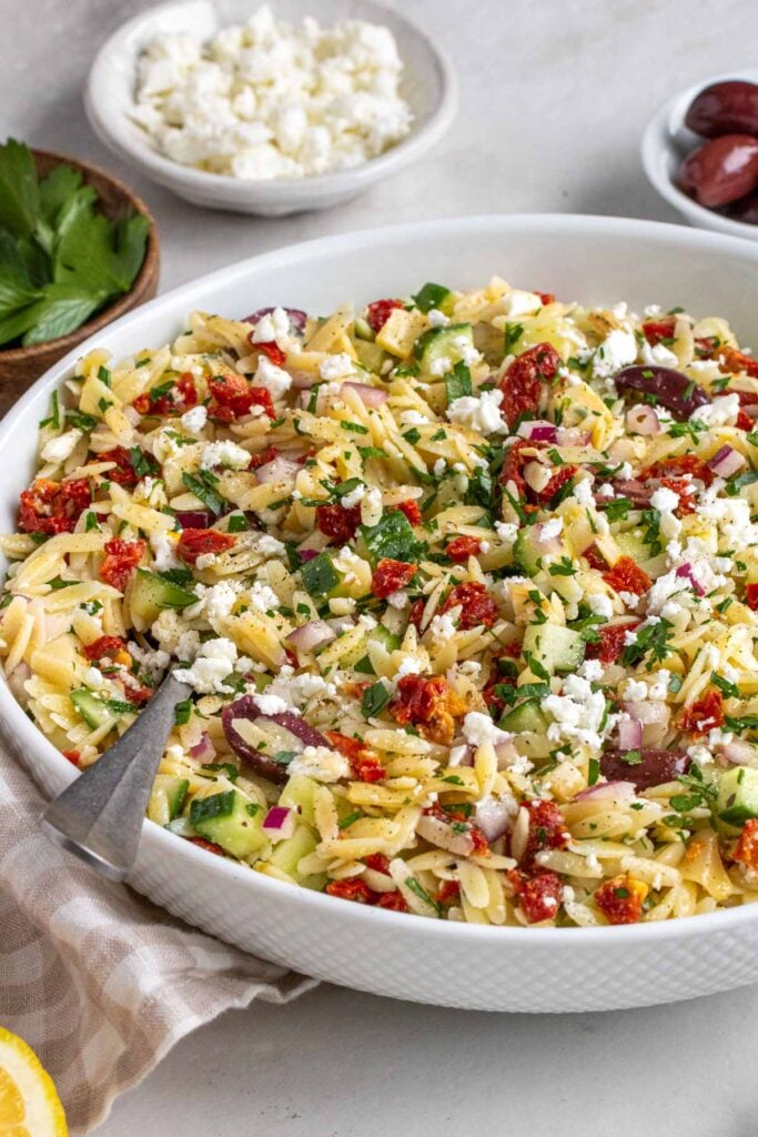 Large white bowl of mediterranean orzo salad with a silver serving spoon on a beige and white checkered tea towel with bowls of parsley, feta cheese, and olives in the background.