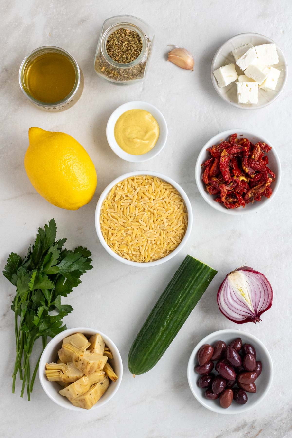 Jar of olive oil, jar of dried oregano, garlic clove, bowl of feta cheese, lemon, bowl of mustard, bowl of sun-dried tomatoes, bowl of orzo, fresh parsley, cucumber, red onion, bowl of kalamata olives, and bowl of artichoke hearts on a white background.