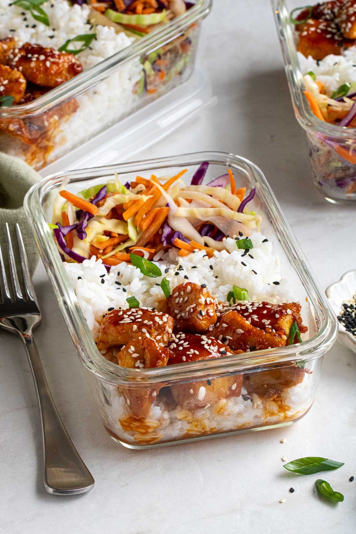 Close up of a sticky chicken rice bowls in a glass meal prep container with a silver fork, green tea towel, sesame seeds, and green onion on the side on a white background.